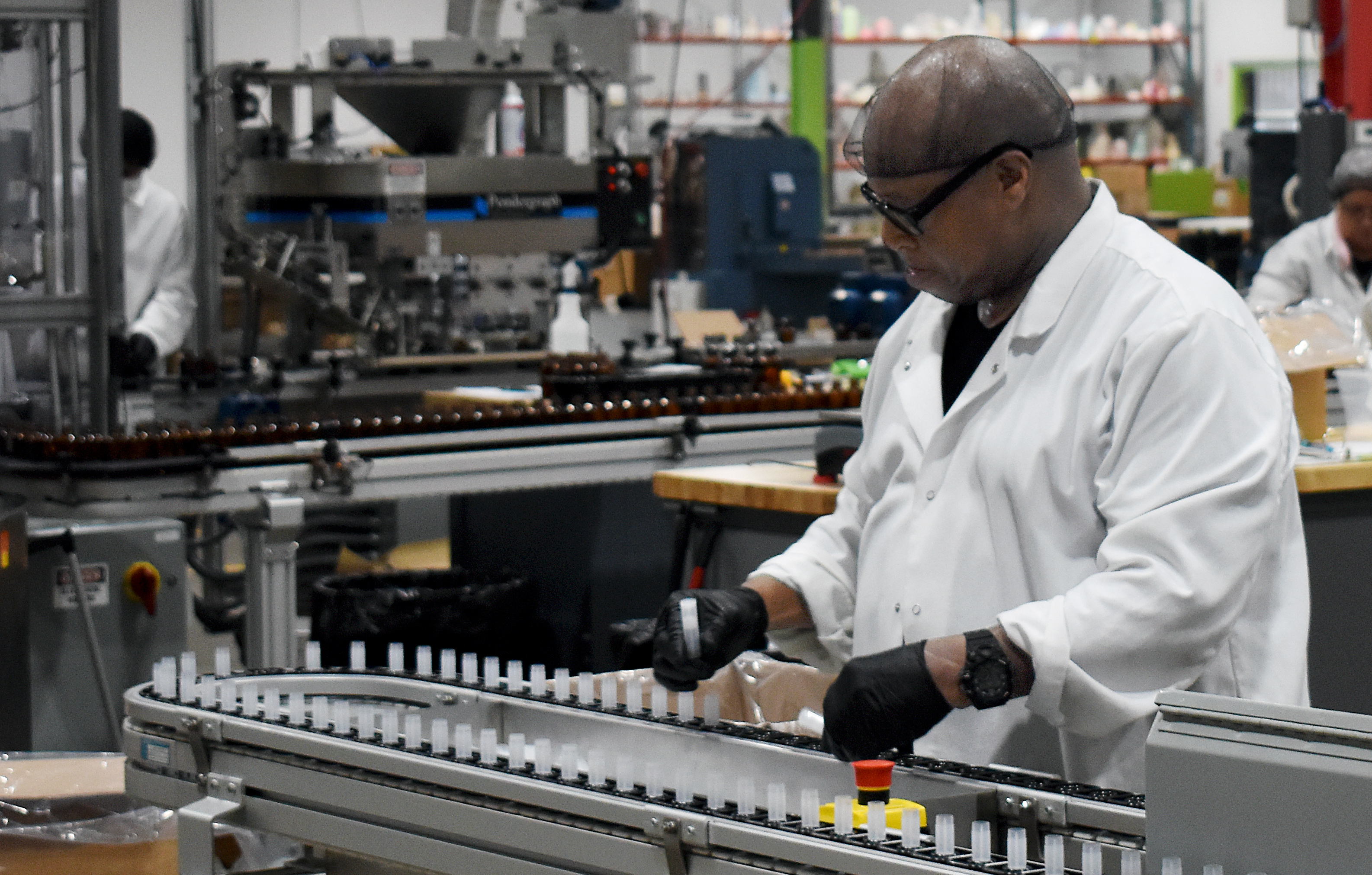 A black man works a lip balm factory line quality checking lip balm tubes on a conveyer. 